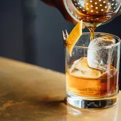 Bursztynowy drink z lodem i skórką pomarańczy, nalewany z shakera. Miękkie, rozmyte tło tworzy przyjemny **bokeh**.