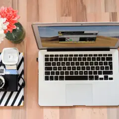 Jak zgrać zdjęcia z aparatu na laptop? Na biurku leży aparat, notatnik, laptop i myszka.