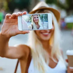 Uśmiechnięta blondynka w kapeluszu robi selfie telefonem, trzymając kawę. Idealny sposób, jak zrobić ładne zdjęcie profilowe.