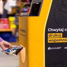Ręka wkłada zdjęcia do automatu, by je wywołać w Empiku. Chwytaj chwile, drukuj wspomnienia.
