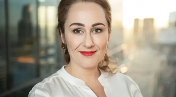 Portret kobiety z rudymi włosami, uśmiechniętej, w białej bluzce. W tle rozmyte miasto.