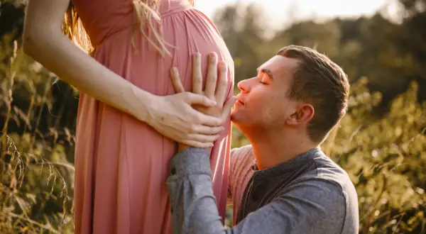 Ciepła sesja ciążowa z mężem. On klęczy i całuje brzuch ukochanej, która delikatnie go obejmuje.