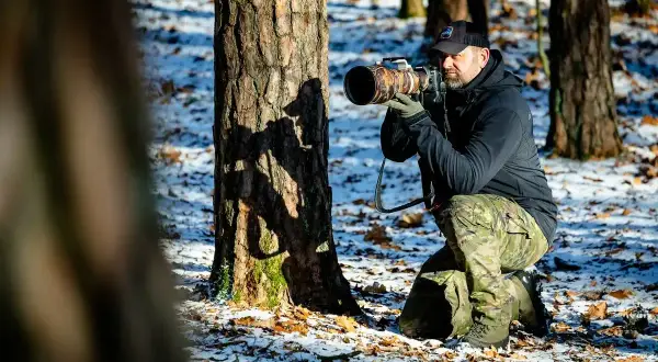 Fotografia przyrody: mężczyzna w kamuflażu kuca przy drzewie, celując profesjonalnym aparatem.