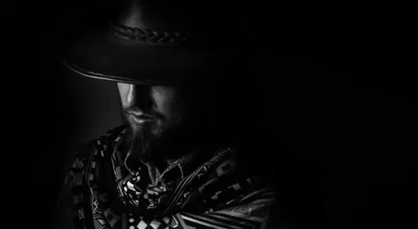 Mężczyzna w kapeluszu i wzorzystym szalu, z twarzą skrytą w cieniu. Dramatyczne chiaroscuro podkreśla tajemniczość postaci.