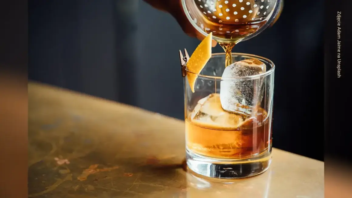 Bursztynowy drink z lodem i skórką pomarańczy, nalewany z shakera. Miękkie, rozmyte tło tworzy przyjemny **bokeh**.
