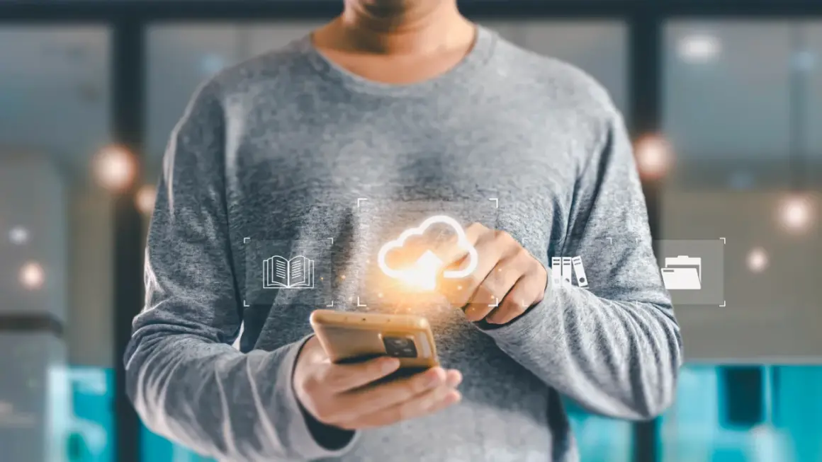 Osoba trzymająca smartfon z ikonami chmury, książki i folderów.
