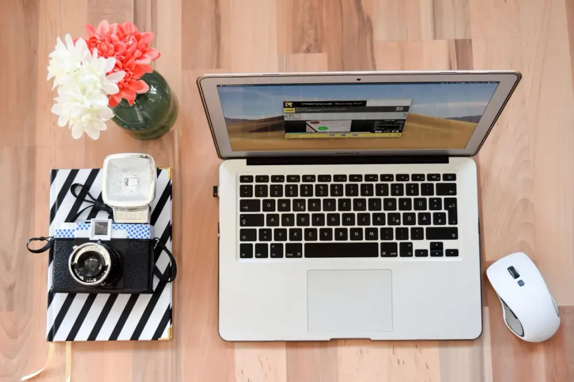 Jak zgrać zdjęcia z aparatu na laptop? Na biurku leży aparat, notatnik, laptop i myszka.